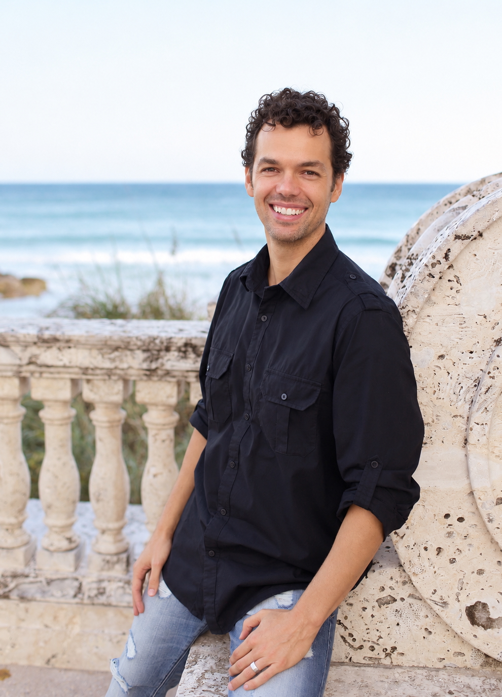 Smiling man in a black button-down shirt leaning on a stone railing with the sea in the background at a coastal location